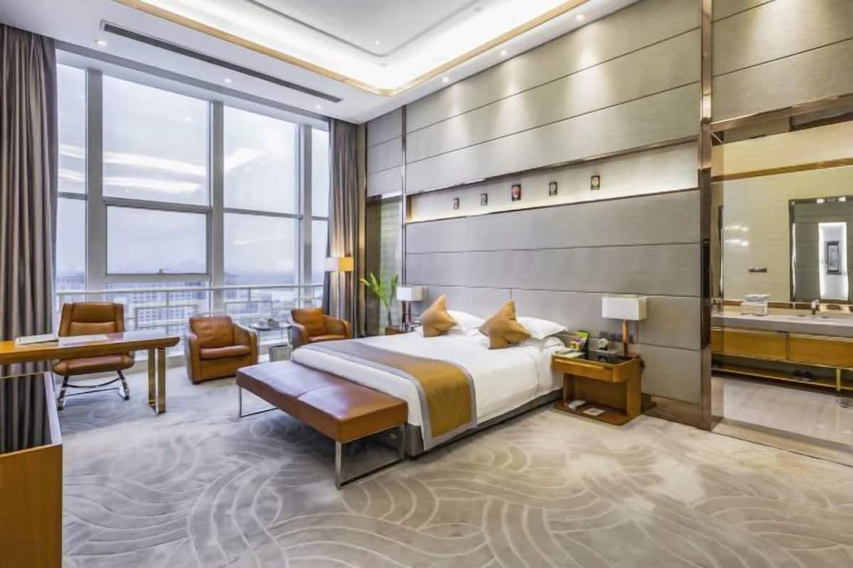 Una habitación de hotel moderna con una cama grande, un escritorio, una silla, un banco, una mesita de noche, un espejo y vistas a la ciudad.