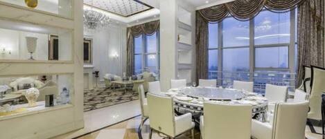 A luxurious dining area with a round table and white chairs, a chandelier, and large windows with curtains.