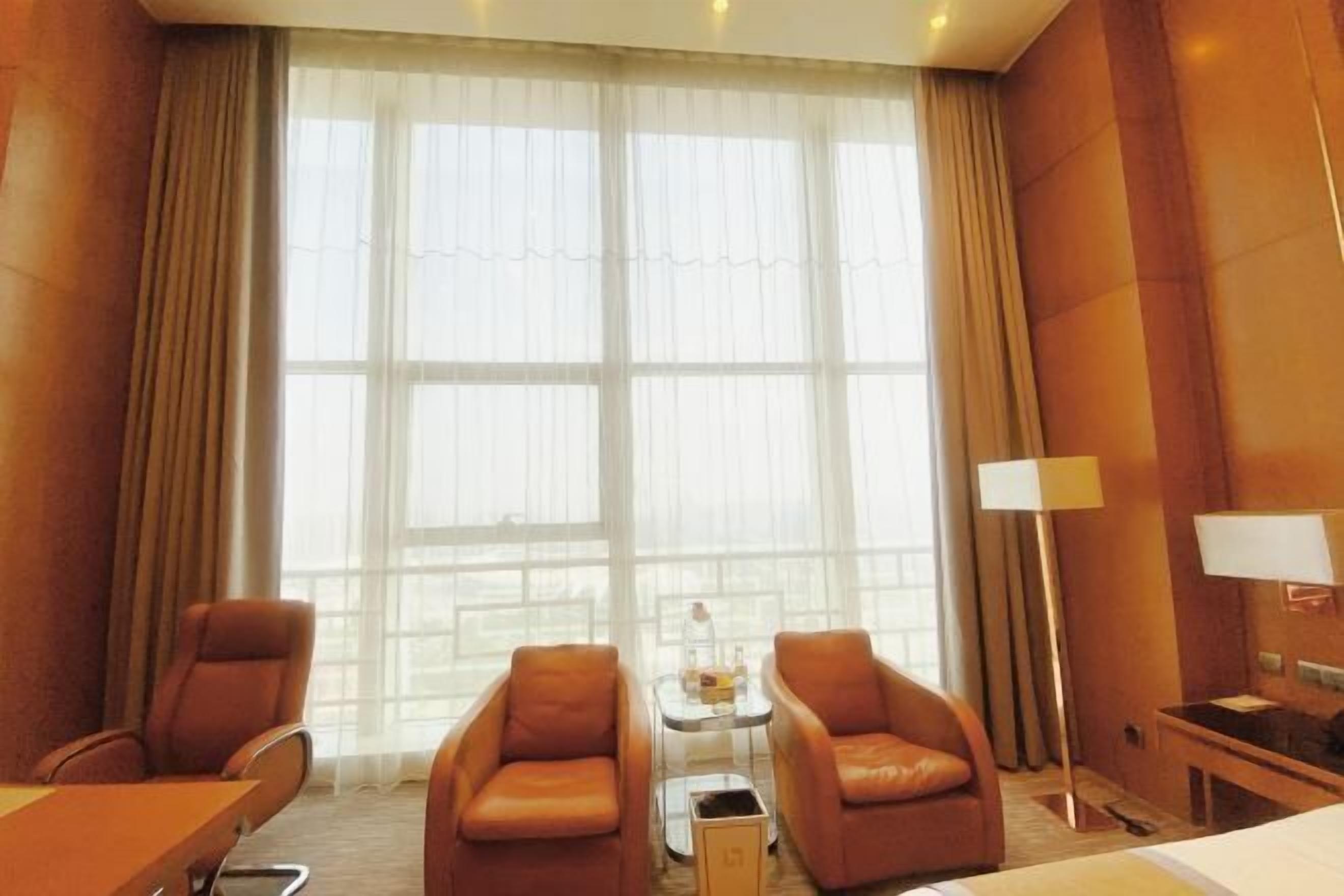 A hotel room with a large window, two leather chairs, a small table, a lamp, and a bed.