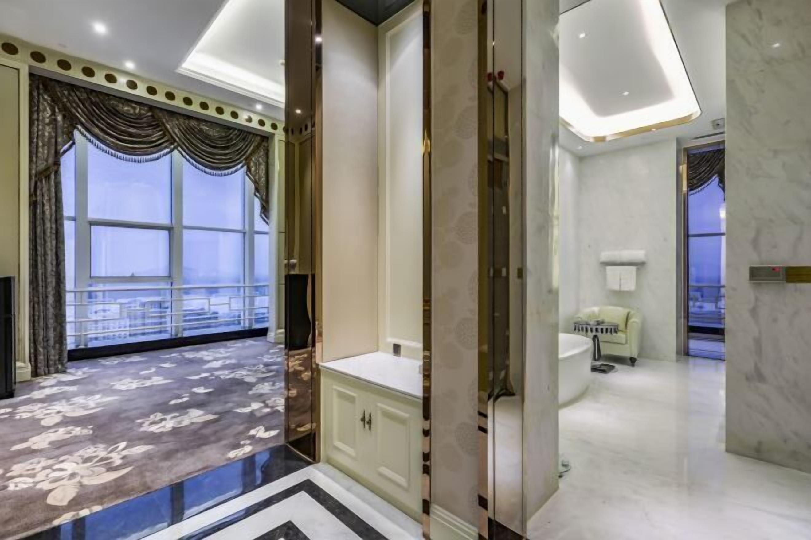 Quarto de hotel luxuoso com uma janela ampla, piso de mármore e banheiro moderno com banheira e vaso sanitário.