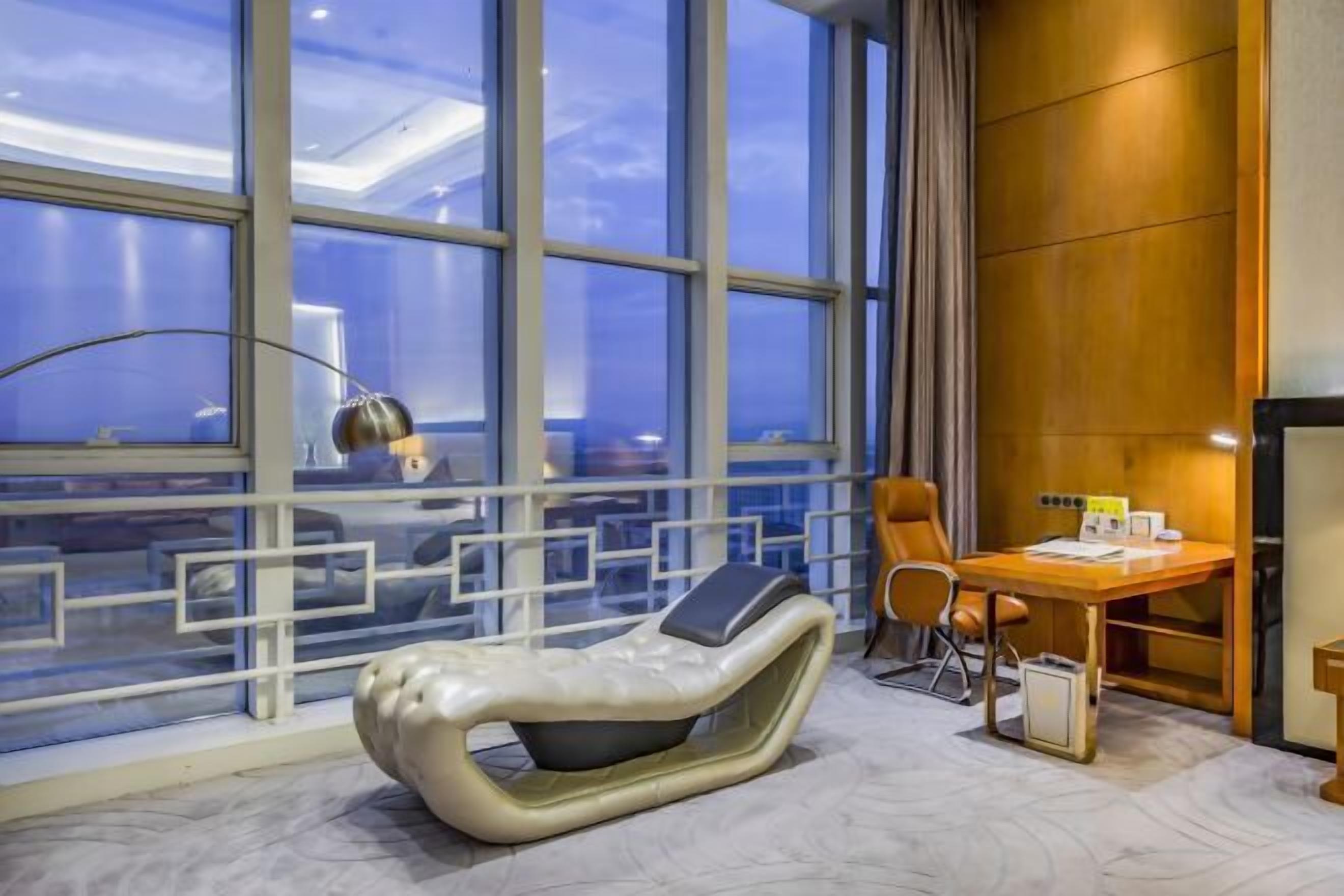 Moderni hotellihuone, jossa on suuret ikkunat, hierontatuoli, työpöytä ja tuoli.
