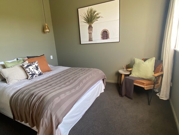 A hotel room with a bed, a chair, a small table with a plant, and a wall-mounted picture.