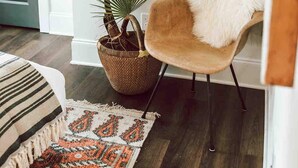 A room with a large mirror, a wooden floor, a brown chair with a white fur throw, a woven basket with plants, and a patterned rug. - Premium 3BR Home by MHM Luxury Properties (Athens)