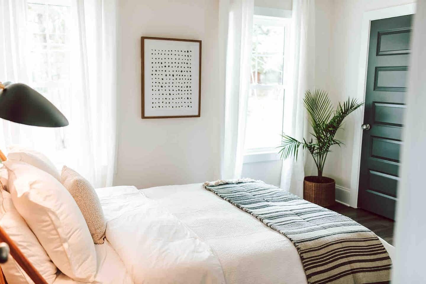 Een slaapkamer met een bed, een kunstwerk in een lijst, een kamerplant en een deur.