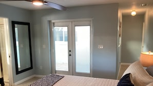 Un dormitorio con cama, ventilador de techo, ventana con persianas, puerta y mesita de noche con lámpara.