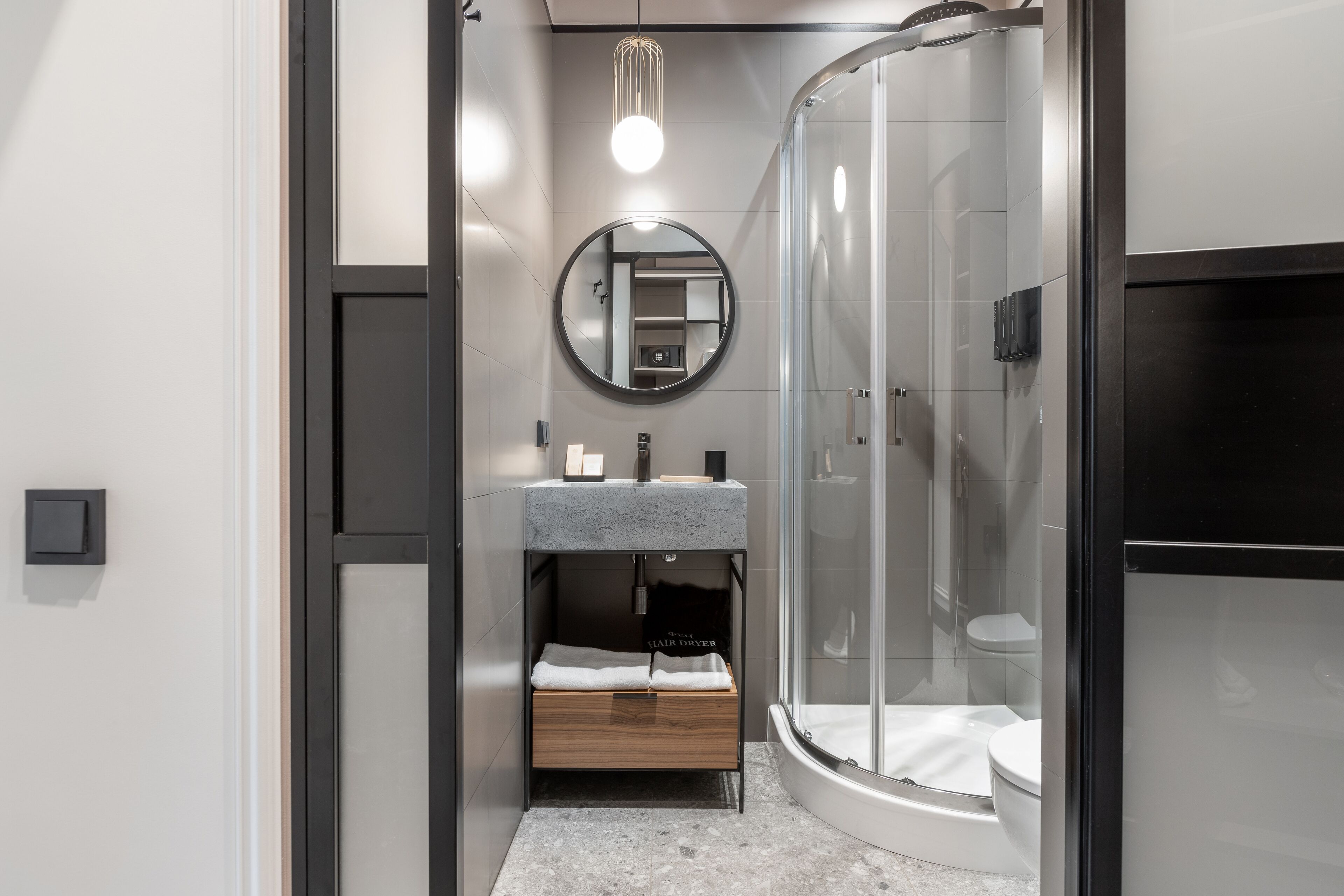 A modern bathroom with a glass shower enclosure, a round mirror, and a built-in wooden storage unit.