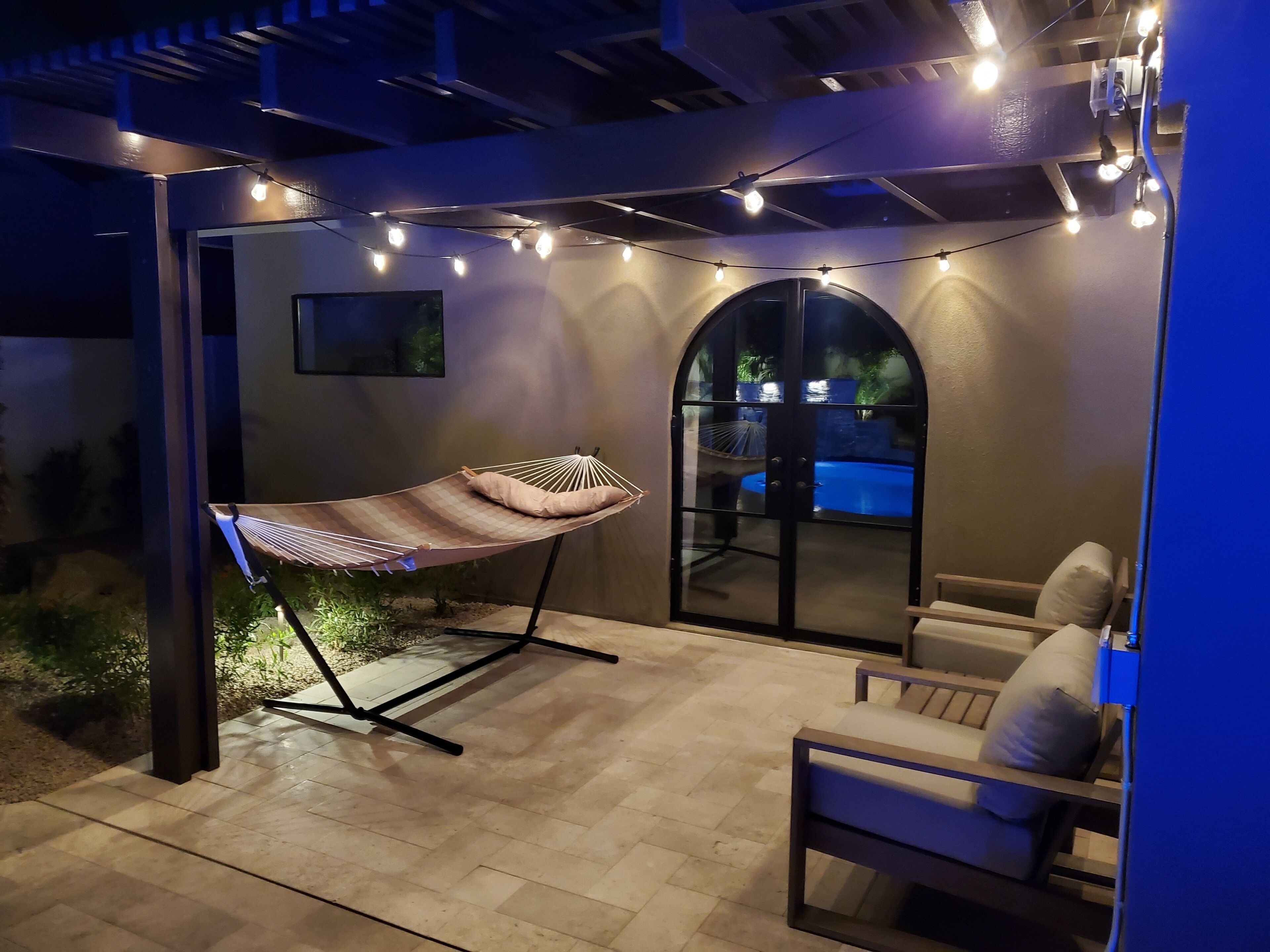 Un patio exterior con una hamaca, una silla, luces colgantes y vista a una piscina a través de un arco.