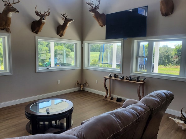 Sala de estar com sofá de couro marrom, mesa de centro de vidro e TV na parede. Há chifres de veado na parede e uma janela com vista para vegetação.