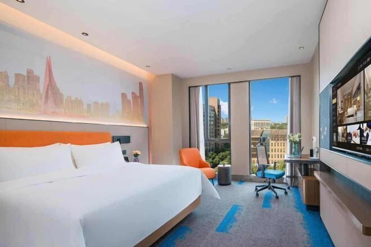 A hotel room with a large bed, a desk, an orange chair, and a large window offering a city view.