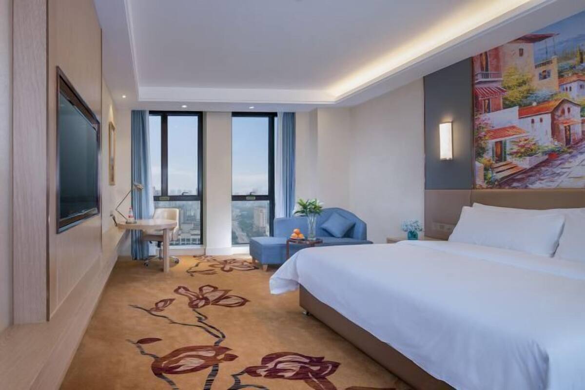 A hotel room with a large bed, a desk, a chair, and a balcony with a city view.