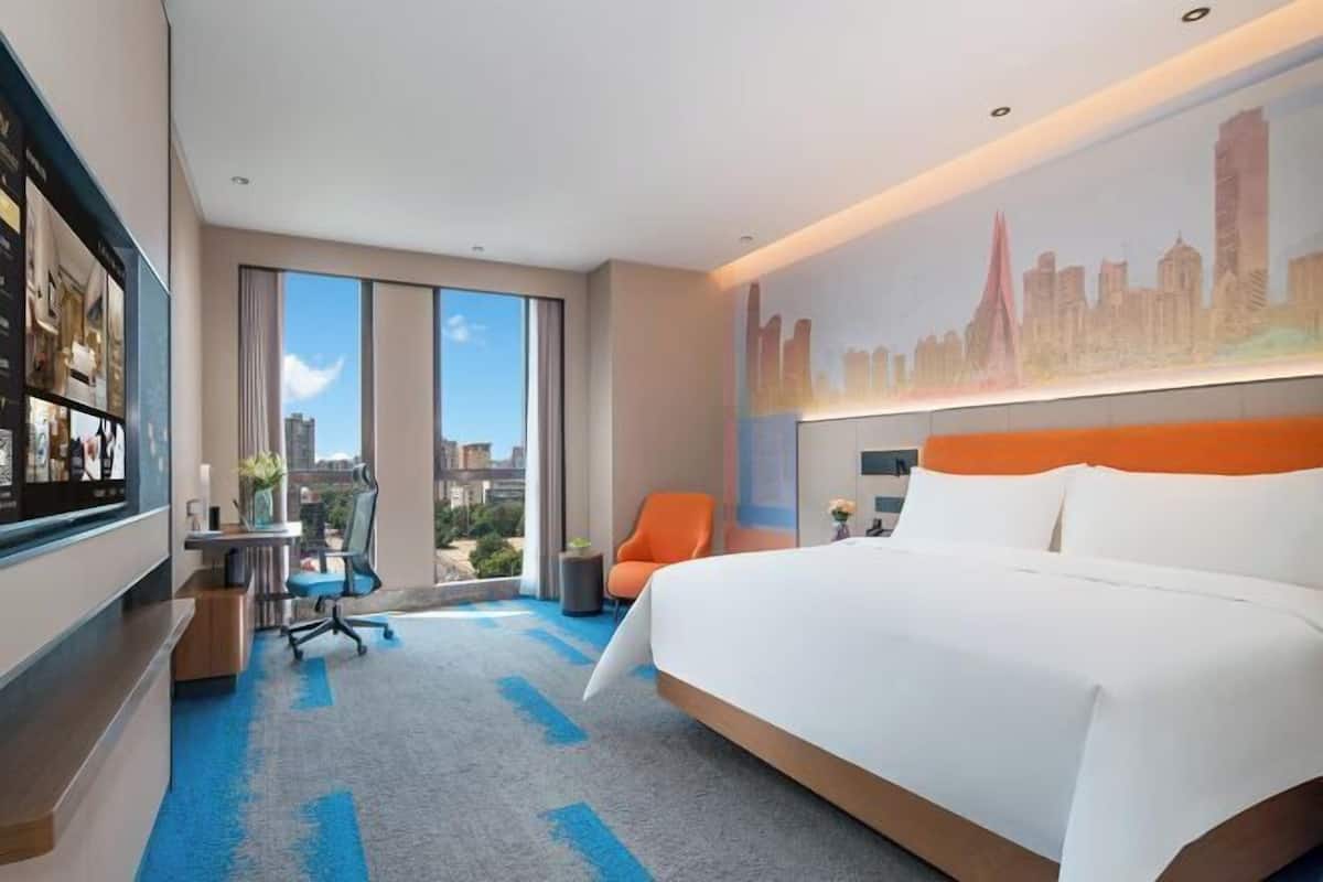 A hotel room with a large bed, a desk with a chair, a TV, and a cityscape mural on the wall.