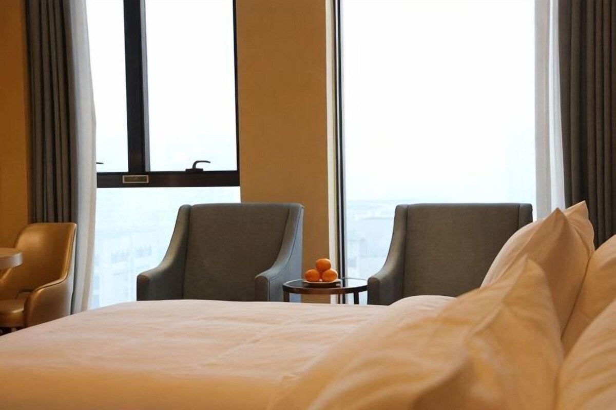 A hotel room with a large bed, two armchairs, a small table with a fruit bowl, and a view through large windows.