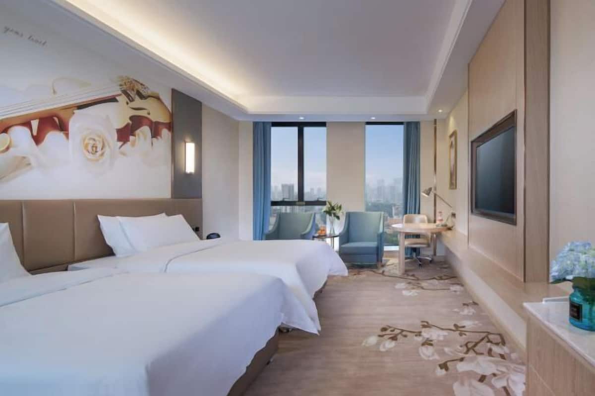 A hotel room with two beds, a desk with a chair, a large window, and a city view.