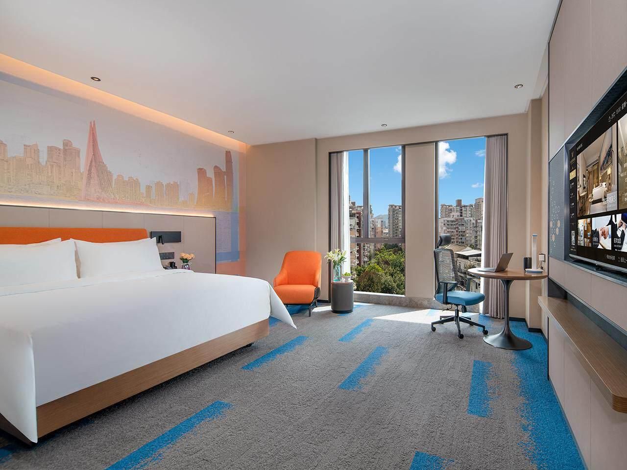 A hotel room with a large bed, a desk with a laptop, a chair, and a view of the city through large windows.