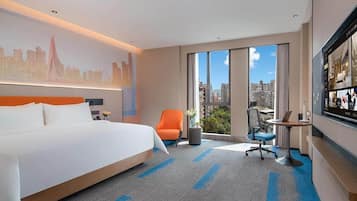 A hotel room with a large bed, a desk with a laptop, a chair, and a view of the city through large windows.