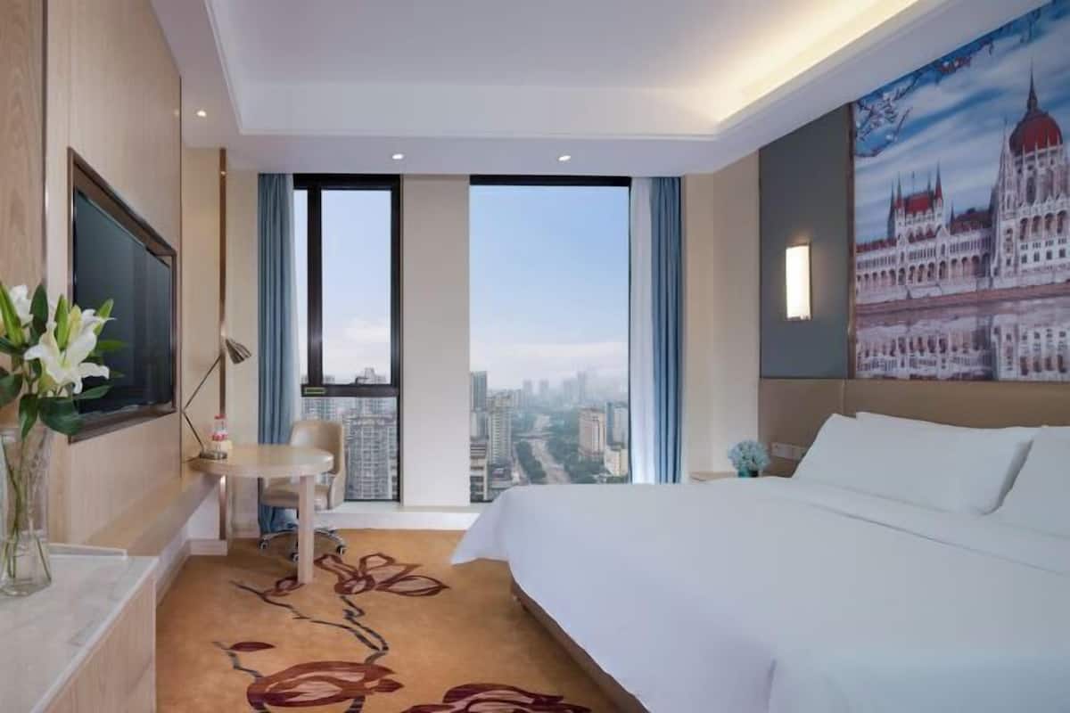 A hotel room with a large bed, a desk, a chair, a TV, and a city view.