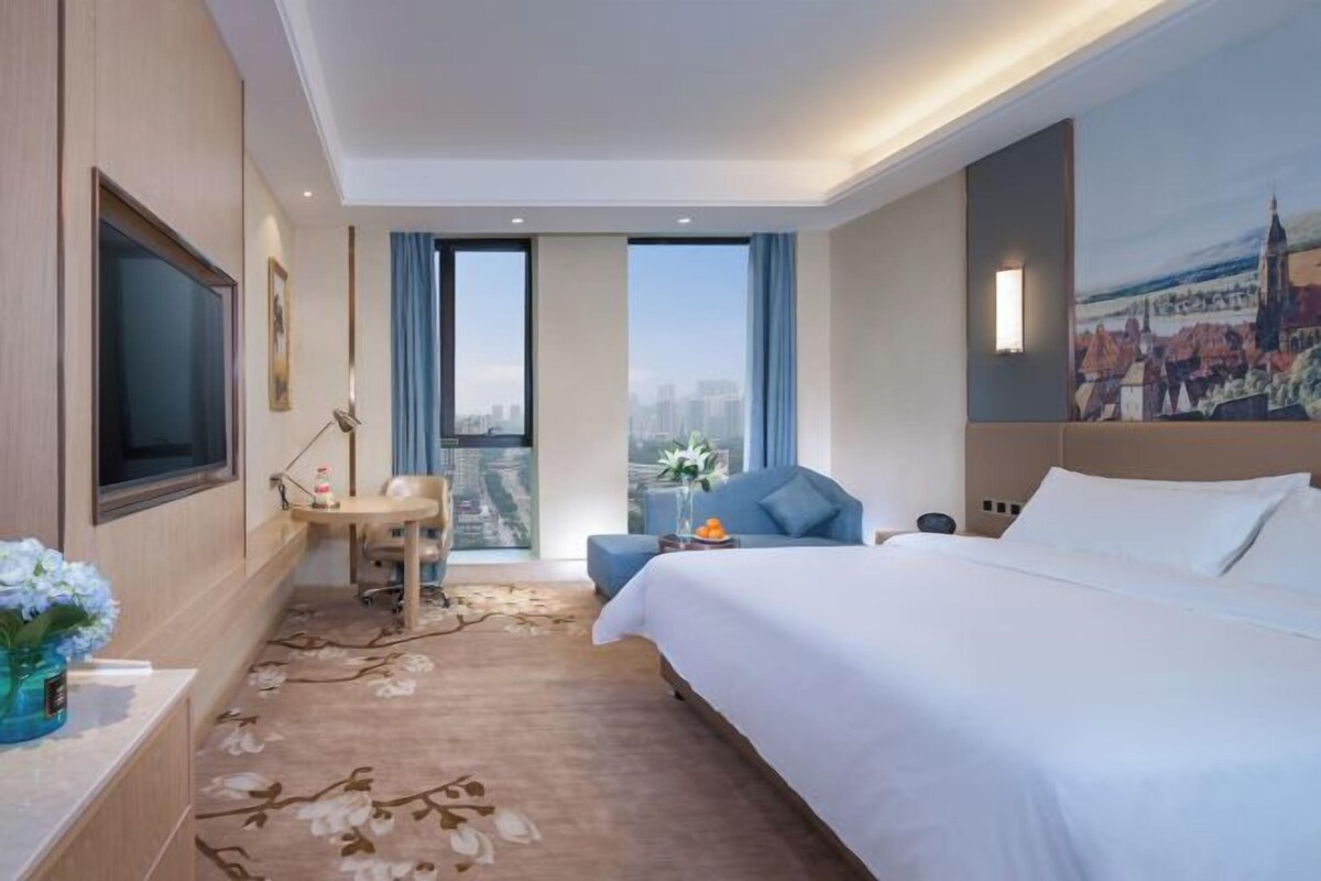 A hotel room with a large bed, a desk, a chair, a TV, and a painting on the wall.