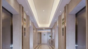 A modern hotel corridor with a central walkway, elevators on either side, and a ceiling with recessed lighting.