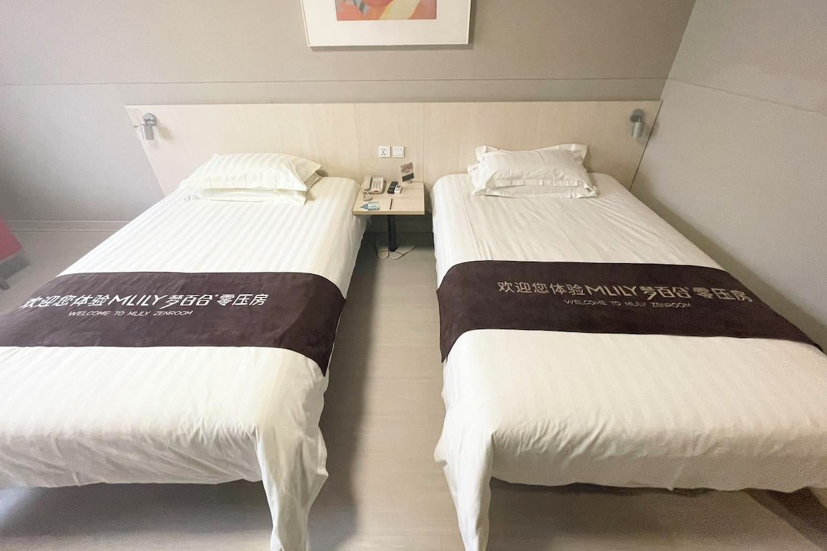 Quarto de hotel com duas camas, cada uma com travesseiro e cabeceira. Há uma pequena mesa entre as camas, com um telefone e um abajur. Uma moldura com uma imagem está pendurada na parede acima das camas.