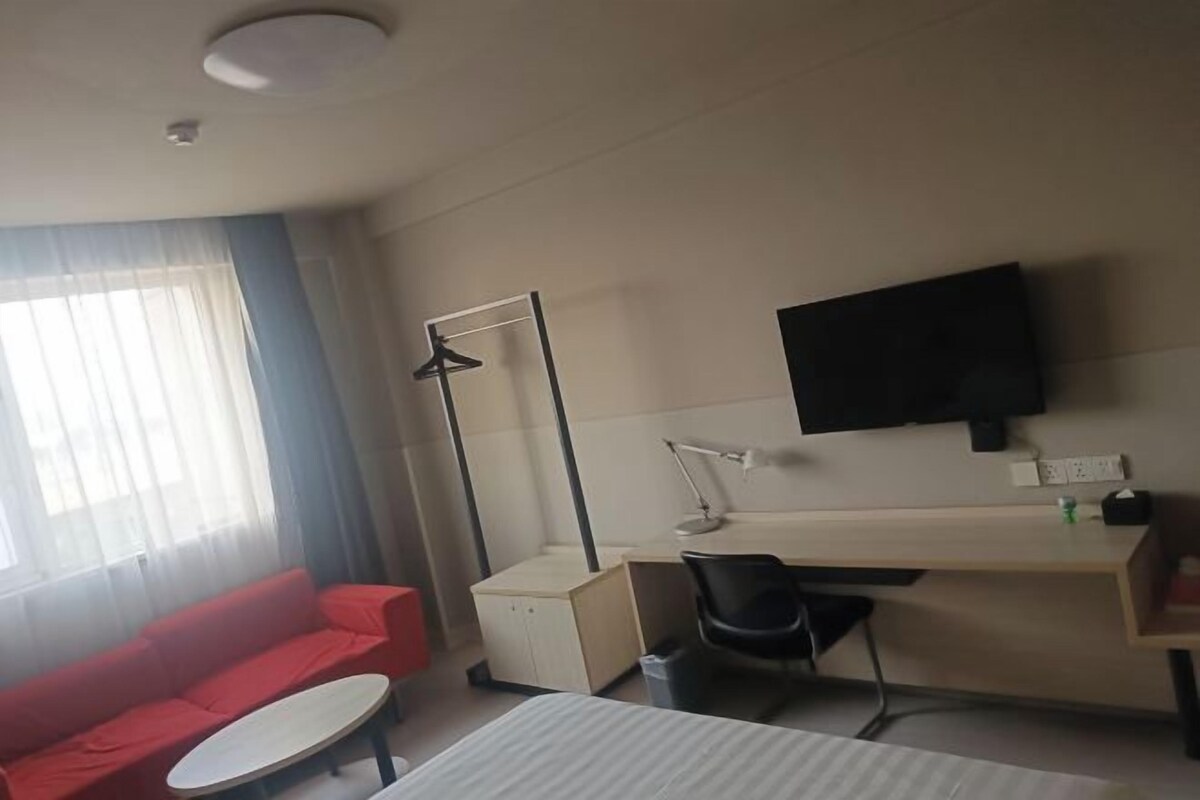 A hotel room with a red sofa, a desk with a chair, a flat-screen TV, and a bedside table.