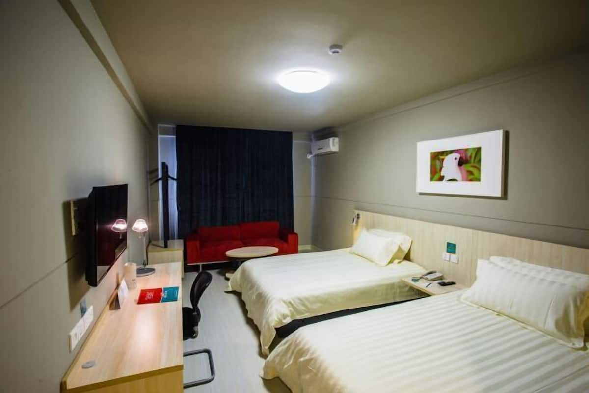 Habitación de hotel con dos camas, un escritorio con lámpara, un sofá rojo, una pequeña mesa redonda y un cuadro en la pared.