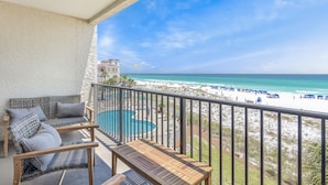 A balcony with wicker furniture and a wooden table overlooking a beach with a pool and umbrellas. - Windancer 307. Gulf Front Condo, Free Beach Service, Heated Pool, Grill. (Miramar Beach)