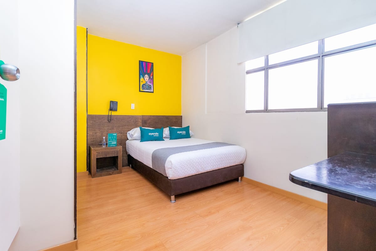 A hotel room with a bed, a nightstand, a small table, a window, and a yellow accent wall.