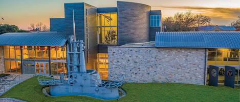 A modern building with a large glass facade, a prominent sculpture in front, and a stone wall with plaques.