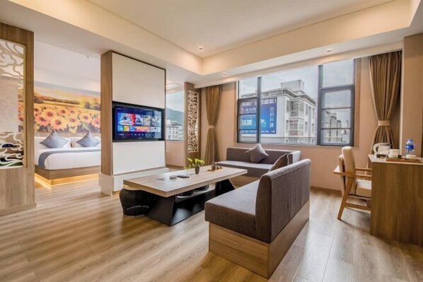 A modern hotel room with a sofa, a coffee table, a TV, and a large window offering a city view.