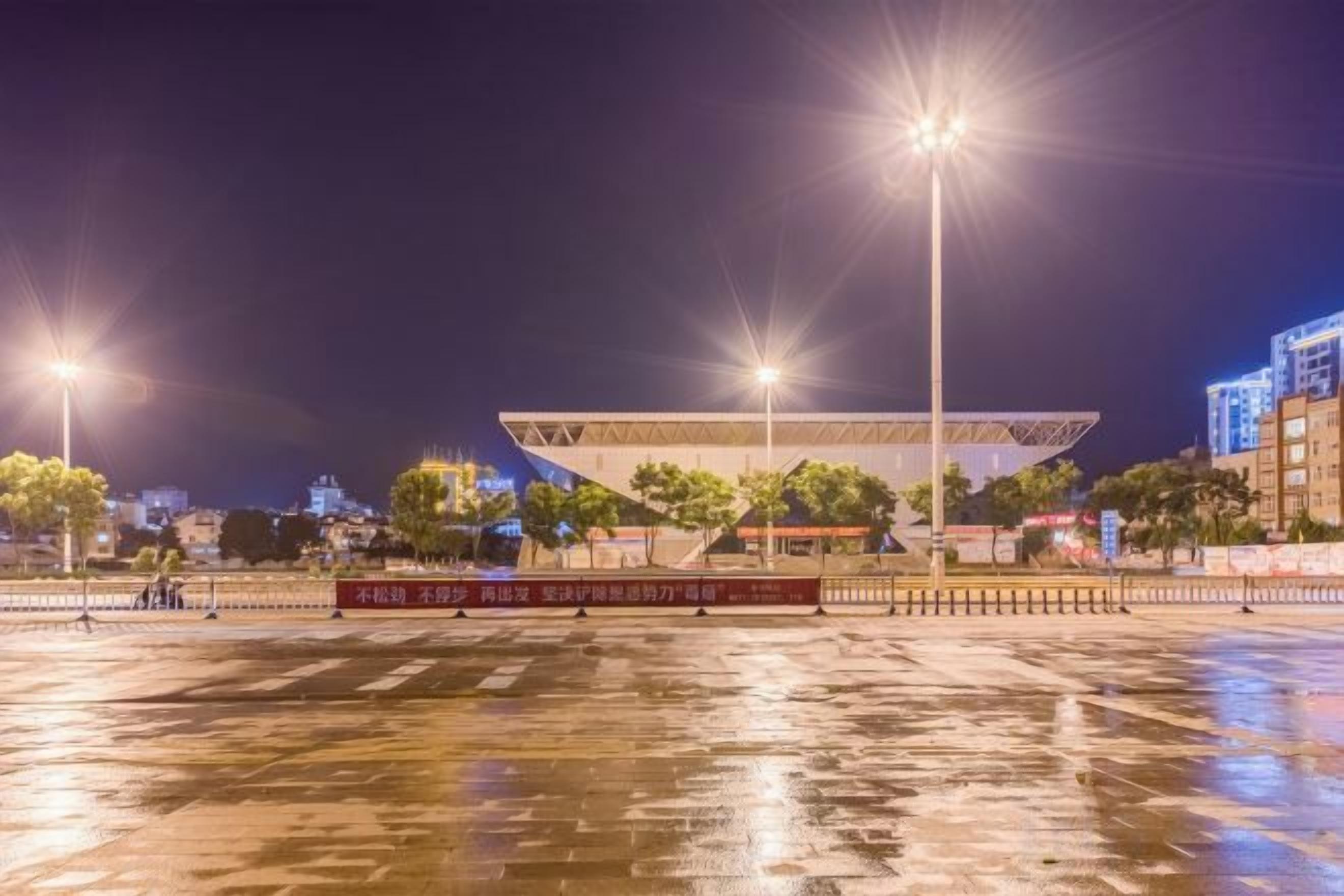 A large, open plaza with a modern building in the background, surrounded by trees and illuminated by street lamps.