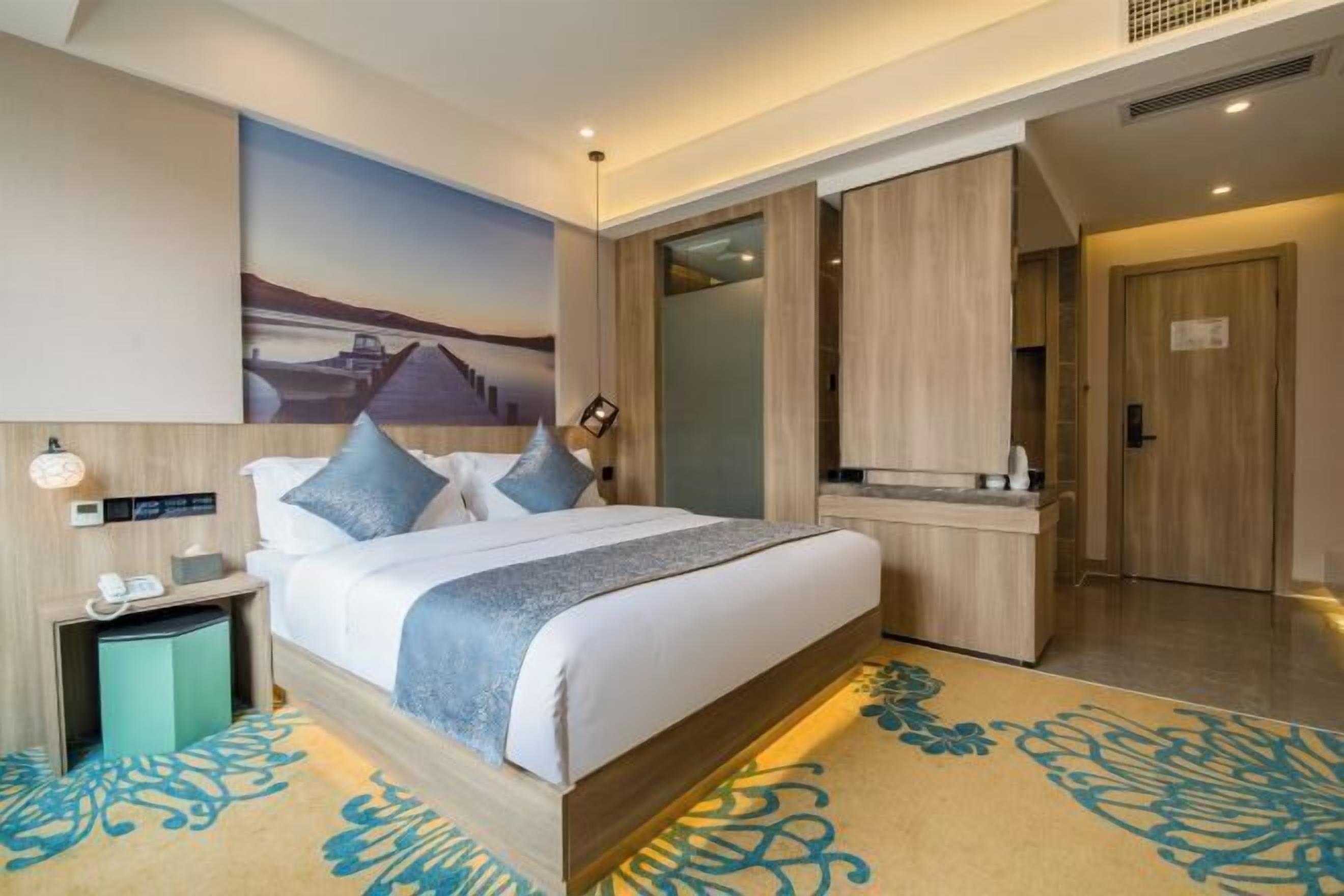 A modern hotel room with a large bed, a bedside table with a lamp, a painting on the wall, and a bathroom door.