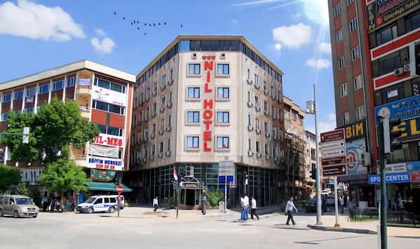 Um edifício em esquina com um destaque para o letreiro "NILHOTEL", rodeado por diversas lojas e um céu claro.