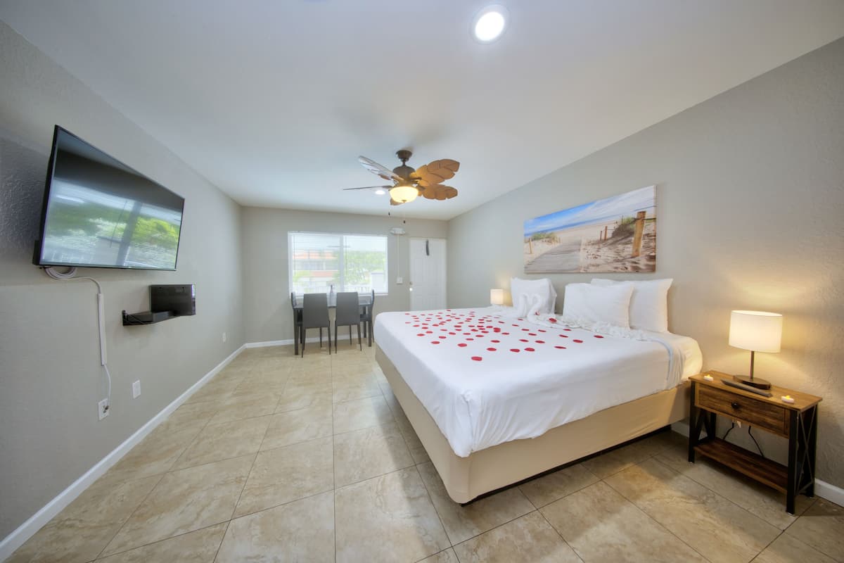 A hotel room with a bed, a desk, a chair, a TV mounted on the wall, a ceiling fan, and a painting on the wall.