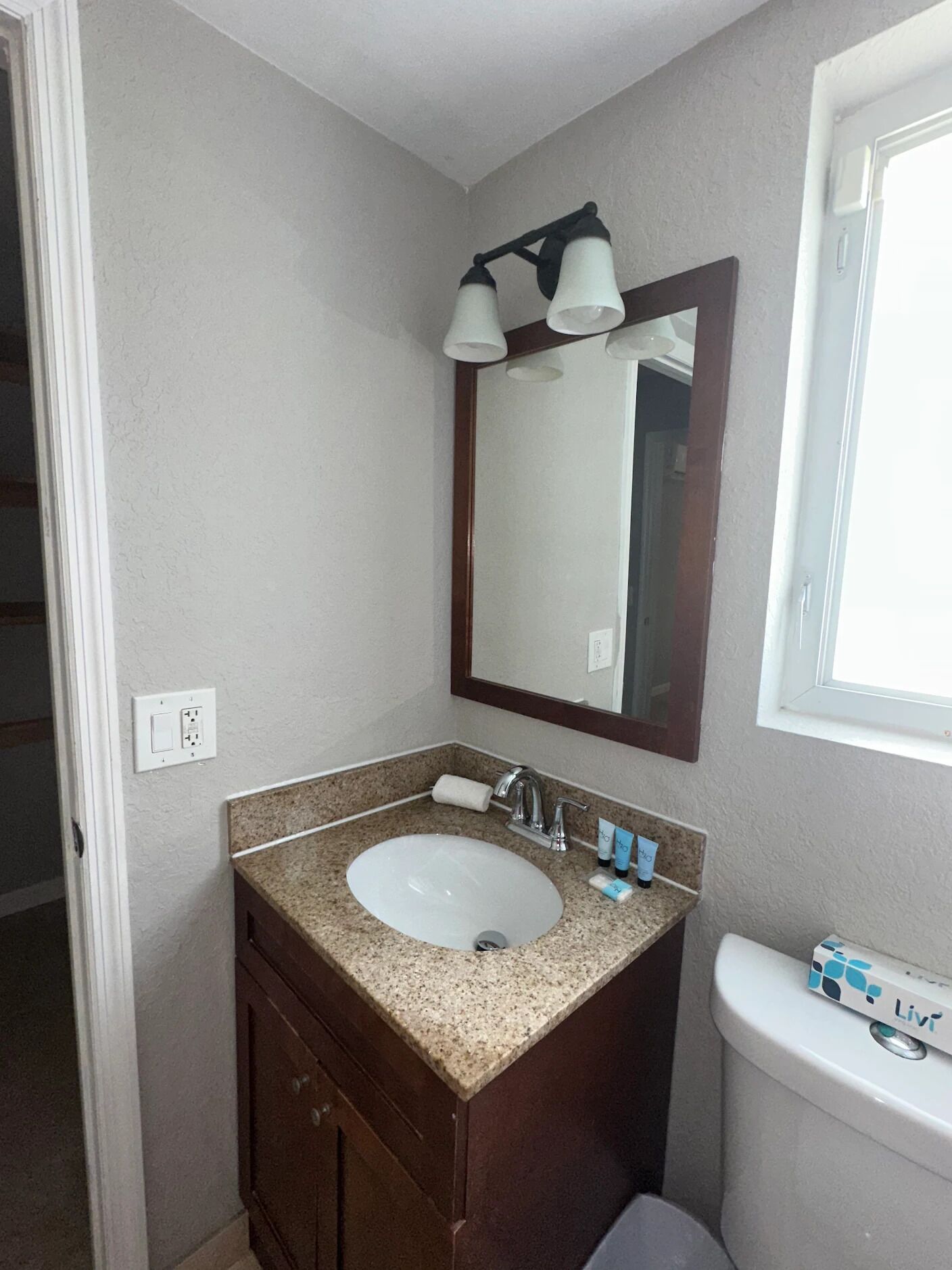 Un baño con lavamanos blanco, tocador de madera oscura, un espejo con tres luces y un inodoro con dispensador de papel higiénico Lix.