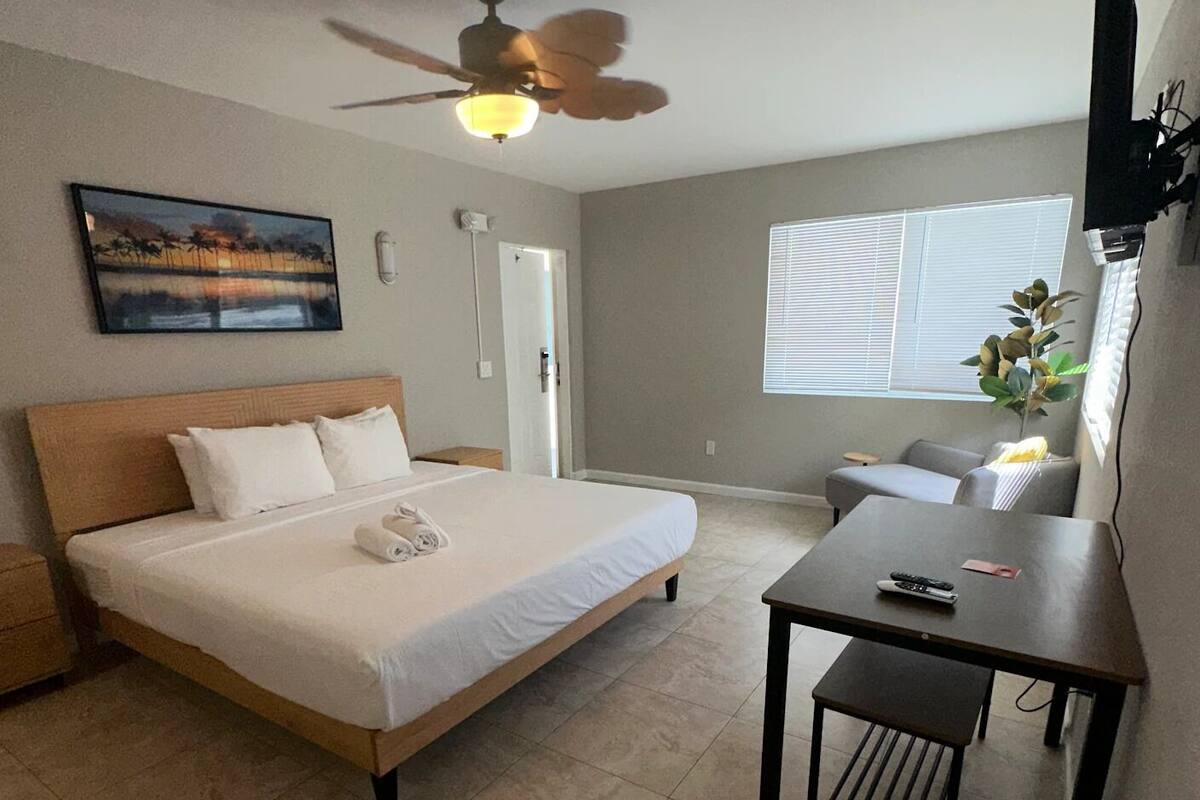 A bedroom with a bed, a desk, a chair, a ceiling fan, a window, and a door.