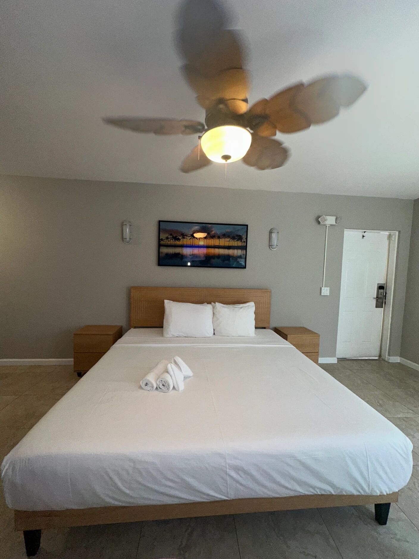 Una habitación de hotel con una cama grande, un ventilador de techo, un cuadro en la pared y una puerta.