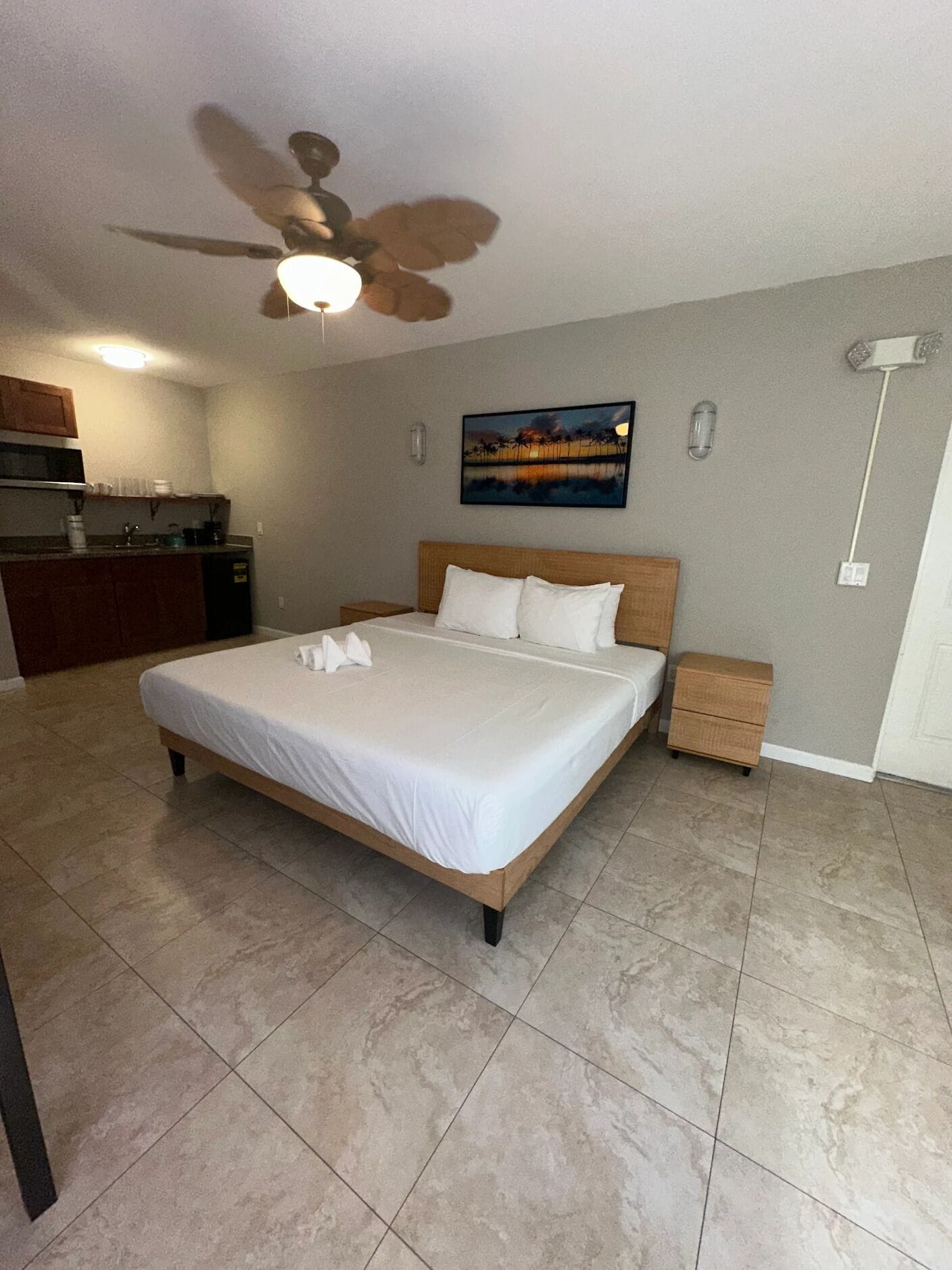 A hotel room with a large bed, a ceiling fan, a kitchen area, and a painting on the wall.