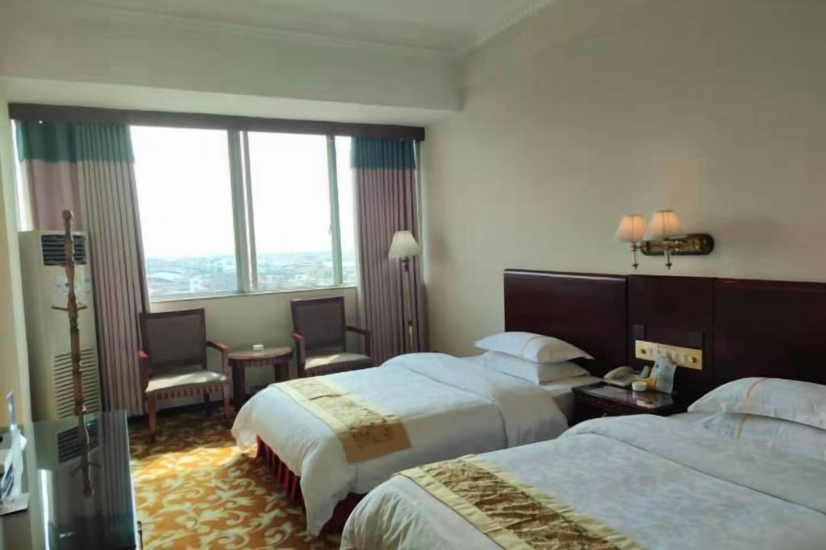 A hotel room with two beds, a desk, a chair, a lamp, and a view of the city.