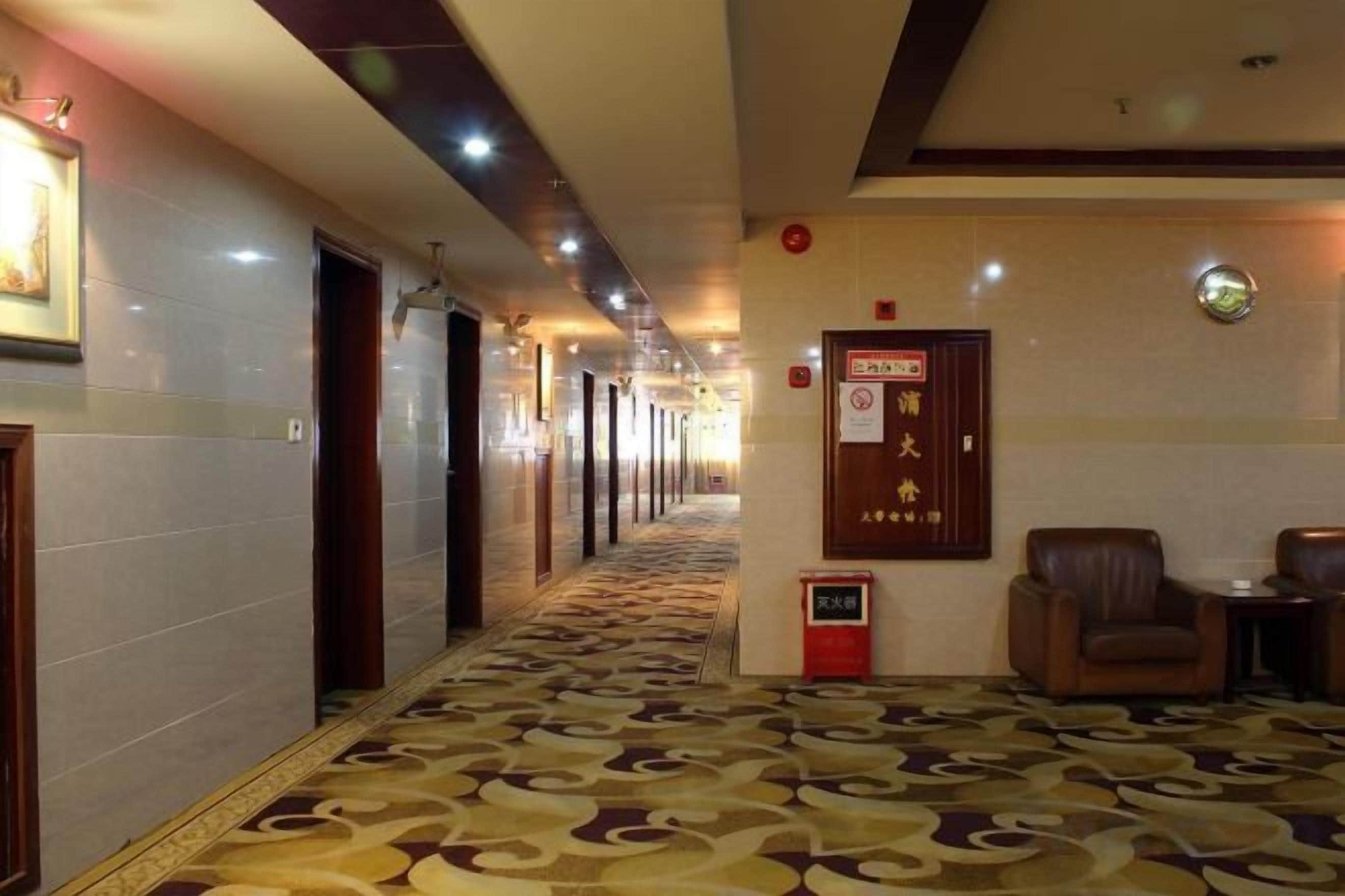 Pasillo de hotel con alfombra estampada, zona de recepción con un cartel y un área de descanso con una silla y una mesa.