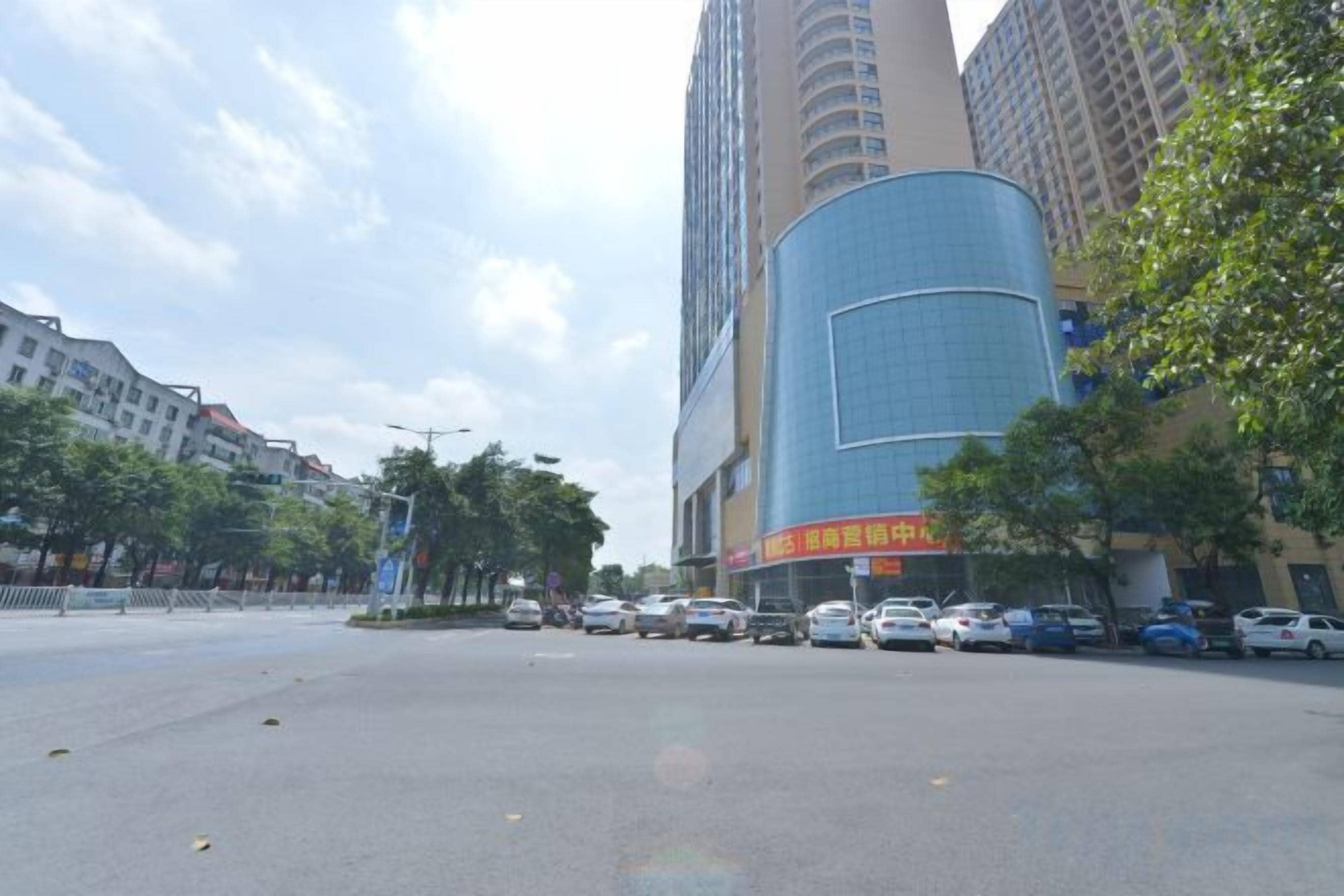 A street view with tall buildings, parked cars, and trees.