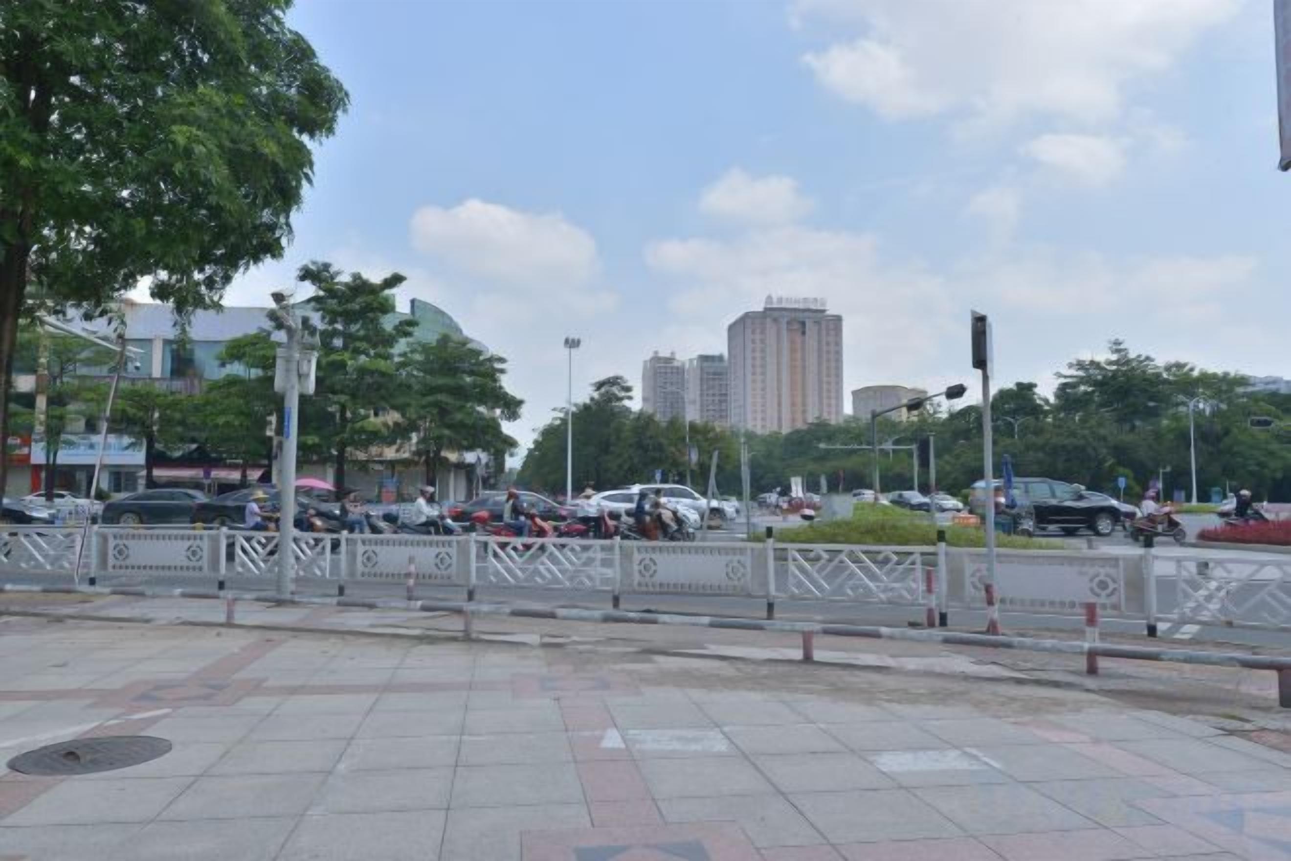 Un área peatonal pavimentada con un cerco blanco, árboles y edificios a la distancia.