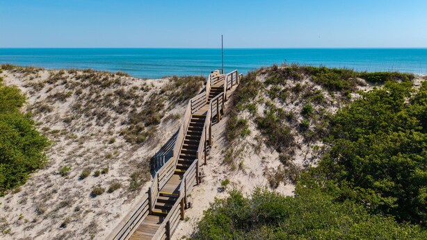 Un escalier en bois qui descend vers une plage, entourée de dunes de sable et d’une végétation clairsemée.