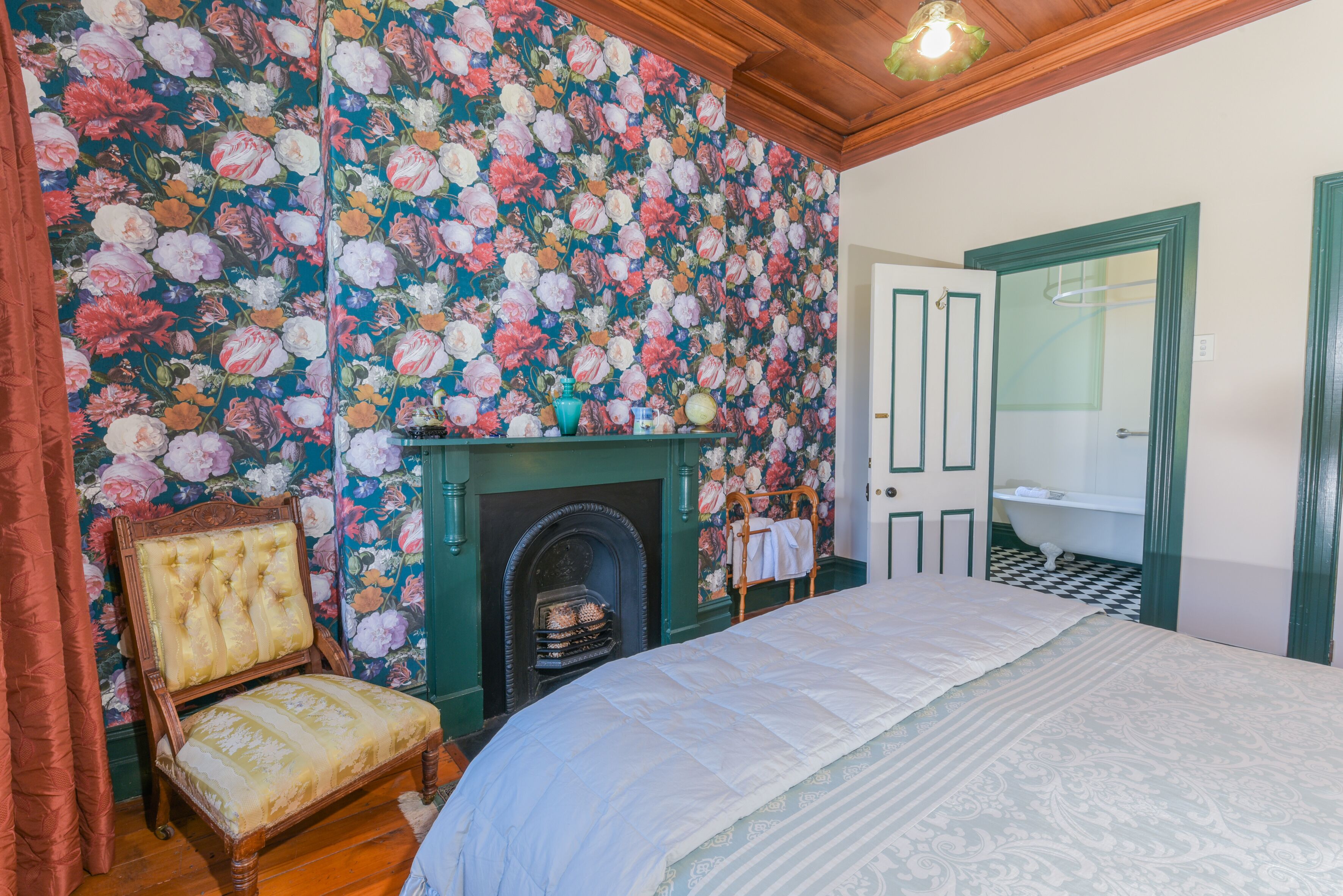 Une chambre à coucher avec un papier peint à motifs floraux, une cheminée, un lit, une chaise et une salle de bain avec un sol à carreaux.
