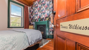 A bedroom with a bed, a fireplace, a floral wallpaper, and a wooden wardrobe with a nameplate. - Dunstan House (Clyde)