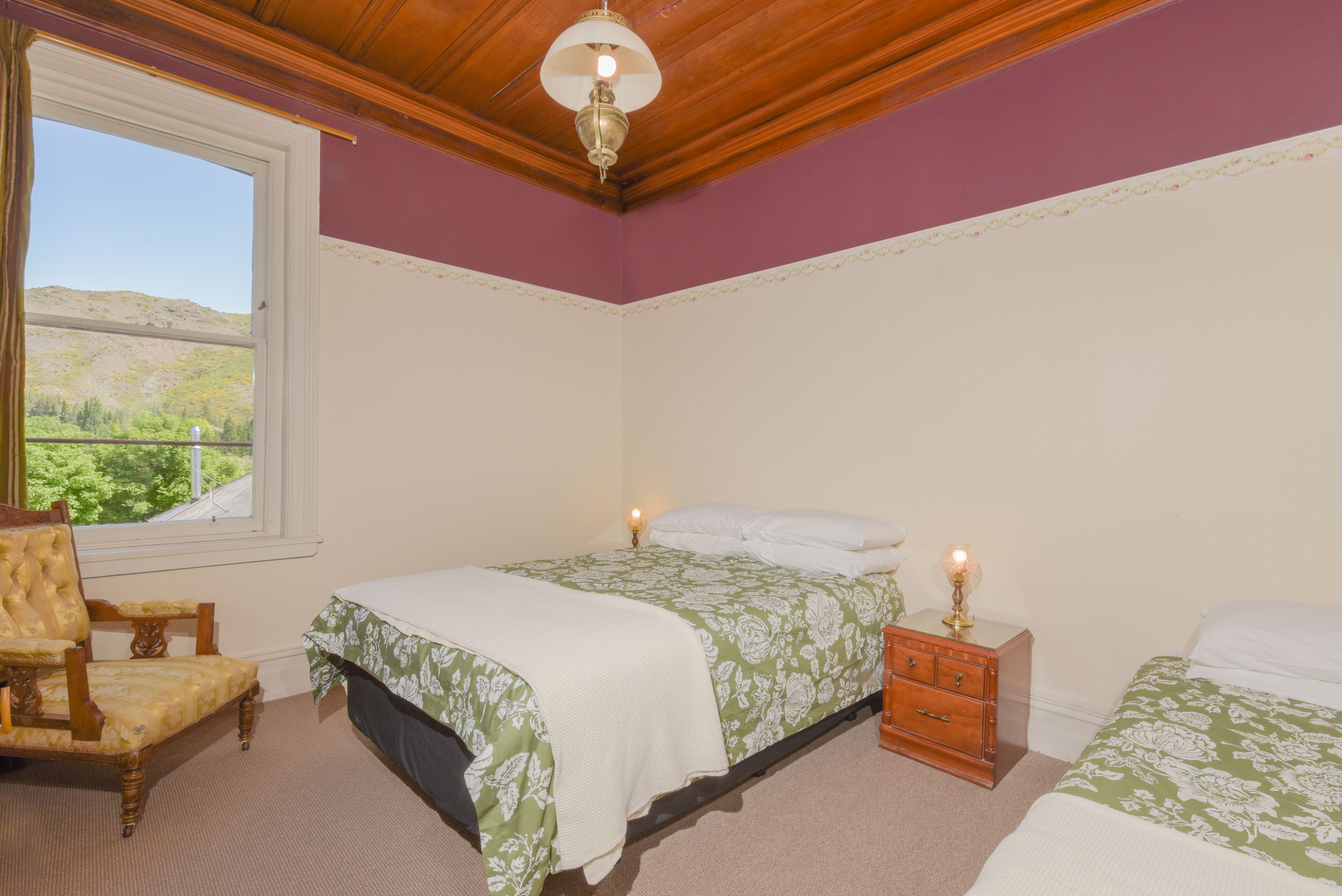 A bedroom with two beds, a wooden chair, a nightstand, and a window with a view of hills.