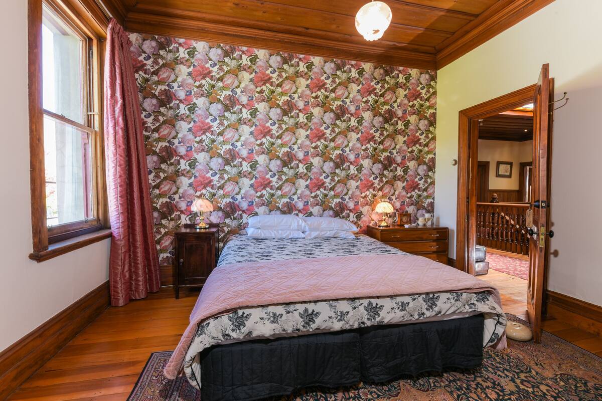 A bedroom with a floral wallpaper, a wooden bed frame, a nightstand with a lamp, and a wooden door leading to another room.