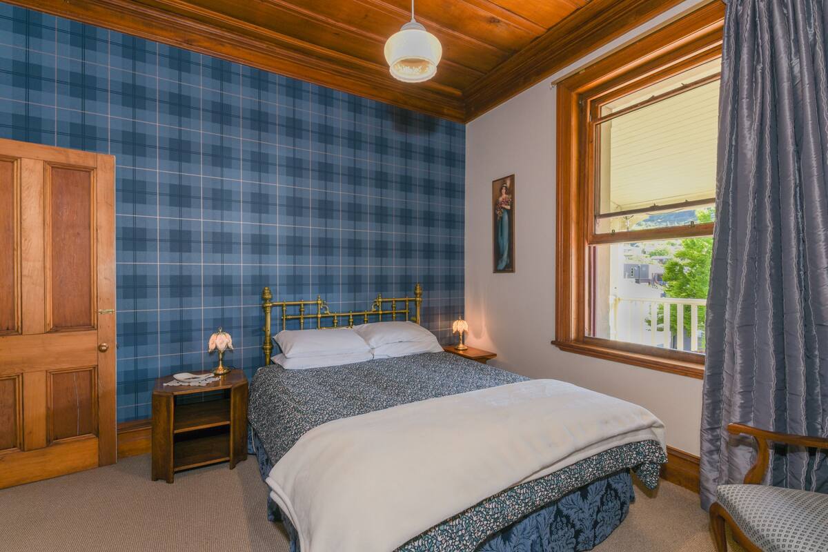A bedroom with a bed, a nightstand, a wooden door, a window with curtains, and a view of the outside.