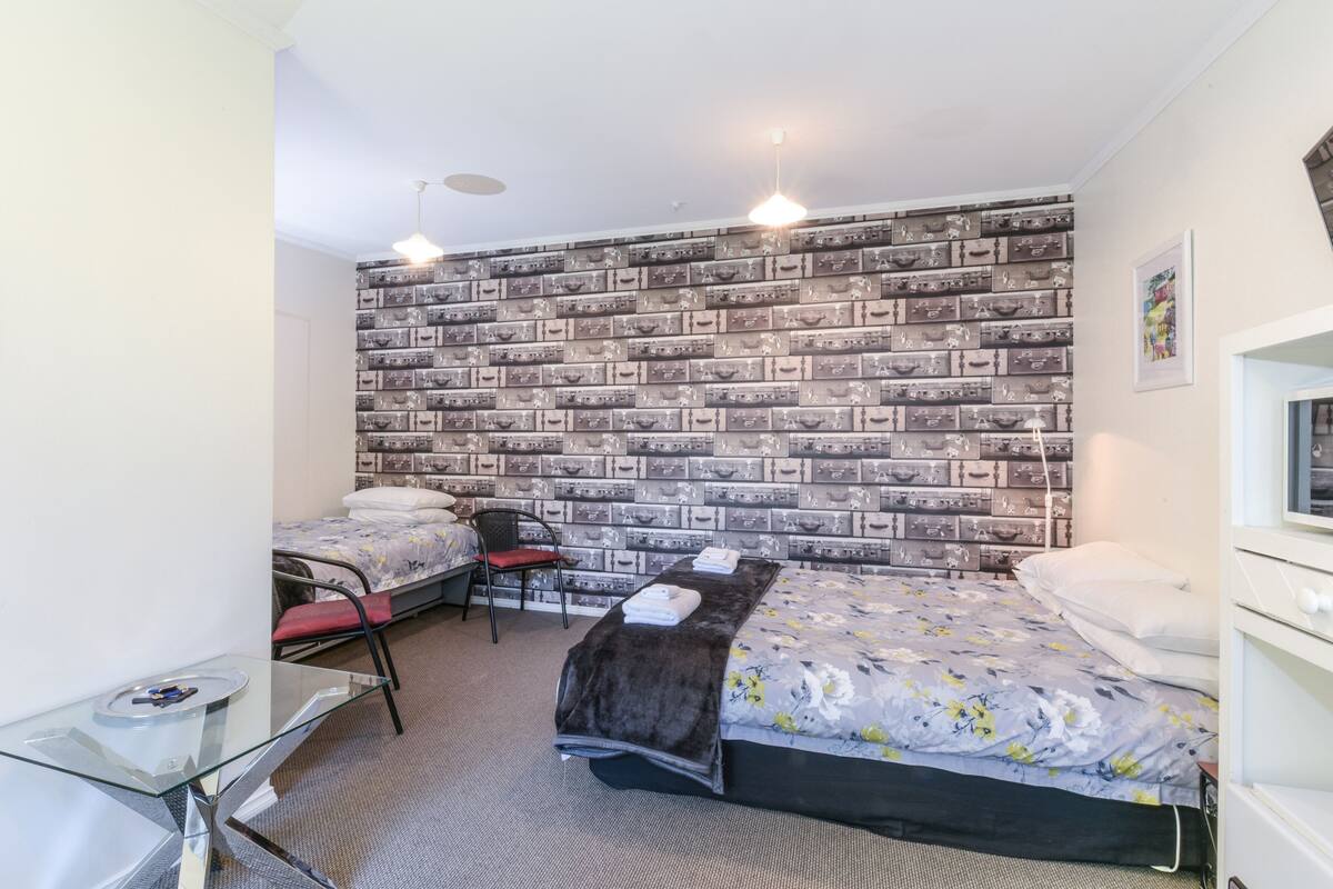 A compact hotel room with a bed, a desk, and a chair. The room has a patterned wall and a ceiling light.