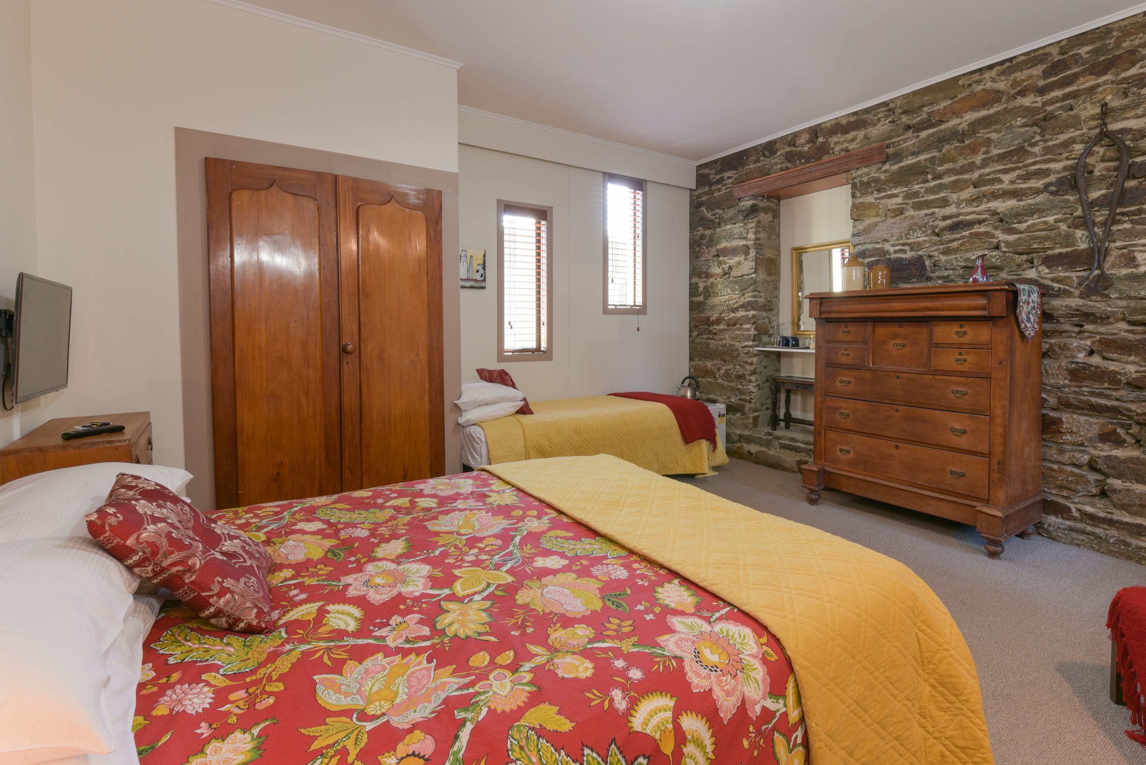 A bedroom with two beds, wooden wardrobe, stone wall, and a dresser.
