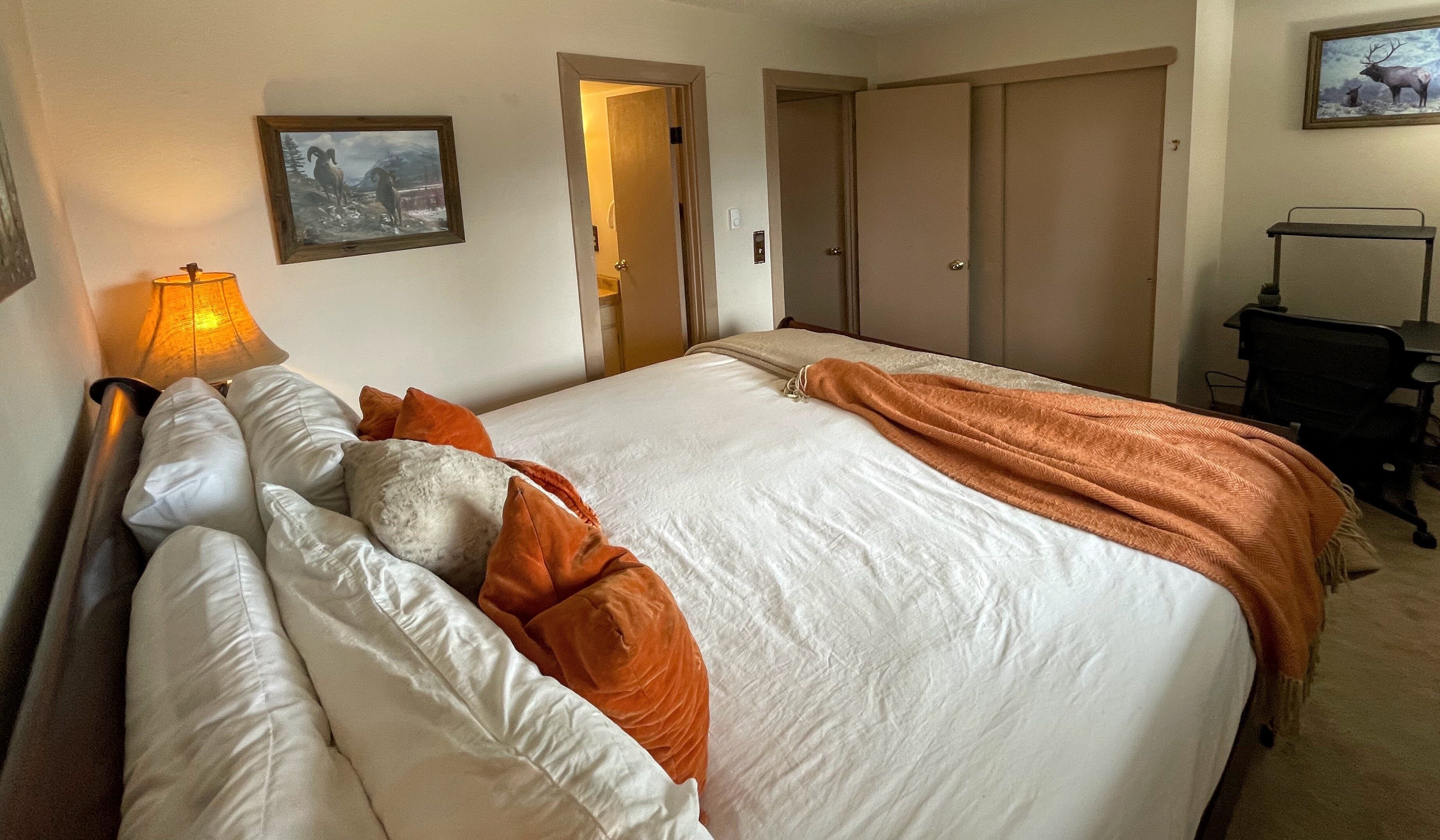 Una cama bien hecha con ropa de cama blanca y naranja, una lámpara y un cuadro en la pared.